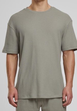 Mann trägt locker sitzendes kurzärmeliges beiges Waffelstrick-Shirt und passende Hose vor einfarbigem Hintergrund.