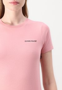 Világos rózsaszín, rövid ujjú póló puha pamutból, kicsi fekete "Guess Jeans" logóval a mellrészen és kerek nyakkivágással.