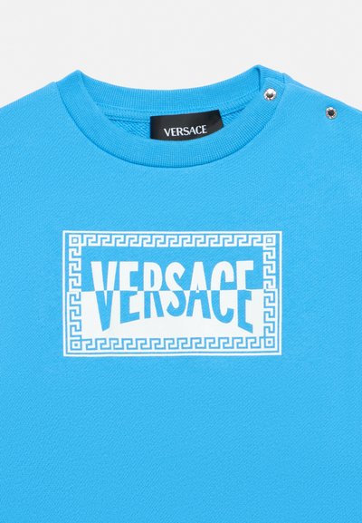 Μπλε μπλούζα με λαιμόκοψη και λευκό λογότυπο Versace στο κέντρο μέσα σε ορθογώνιο πλαίσιο με μοτίβο ελληνικού κλειδιού και δύο ασημένια κουμπιά στον ώμο.