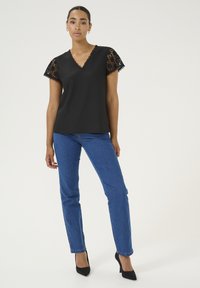 Schwarzes Kurzarmoberteil mit Spitzenakzenten, V-Ausschnitt, kombiniert mit blauen gerade geschnittenen Jeans und schwarzen High Heels vor einem neutralen Hintergrund.
