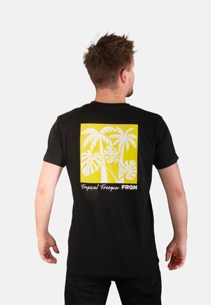 Homme aux cheveux courts marron portant un t-shirt noir avec un carré jaune et un motif de plante tropicale blanche dans le dos.
