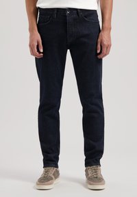 Vaqueros de mezclilla azul oscuro con corte recto, cinco bolsillos y cierre de botón. Llevados con zapatillas gris claro.