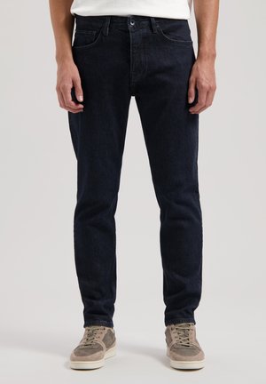 Jeans de denim albastru închis cu o croială dreaptă, cinci buzunare și închidere cu nasturi. Purtați cu adidași gri deschis.