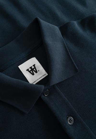 Polo shirt di un blu navy scuro con un tessuto testurizzato, chiusura con bottoni e un'etichetta marchiata cucita al collo. Finitura del colletto liscia.