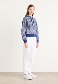 Blå mönstrad bomberjacka med dragkedja fram, ribbade ärmslut och matchande midja, tillsammans med vita sweatpants och vita sneakers.
