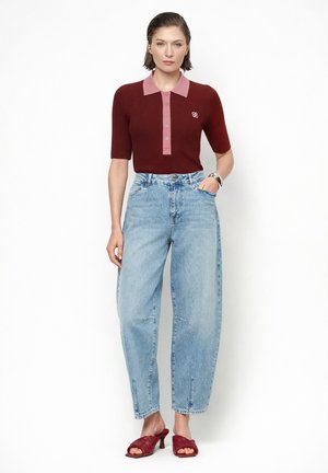 Vrouw draagt een maroon kortärmelig poloshirt, lichtblauwe high-waisted jeans, rode sandalen met hak en een polshorloge, staat met één hand in haar zak.