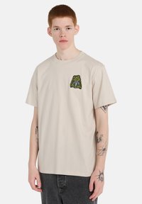 Μπεζ βαμβακερό T-shirt με σχέδιο βατράχου σε πράσινο και μαύρο χρώμα στο αριστερό στήθος. Το πουκάμισο έχει κλασική εφαρμογή και κοντά μανίκια.