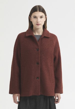 Femme aux cheveux bruns lisses porte un manteau en laine rouge foncé boutonné et une robe gris foncé sur un fond clair uni.