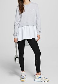 Donna che indossa un felpa grigia sopra una tunica bianca, leggings neri e sneakers bianche multicolore, con una borsa argentata al suo fianco.