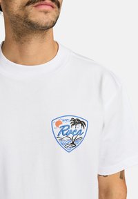 Hvid bomulds-T-shirt med et bl�åt og orange broderet logo, der viser en sol, palmer og bølger, vist fremtrædende på brystet.