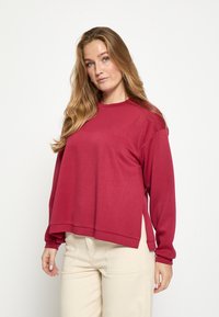 Pikkade varrukatega punane t-särk, mille lõige on lõdvestunud, ümara kaelusega ja külgedel lõhikuga, mis on kombineeritud heledate pükstega. Pehme tekstuur.