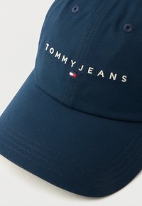 Ναυτικό μπλε καπέλο μπέιζμπολ με κεντημένο λογότυπο "TOMMY JEANS", με μικρή λεπτομέρεια σημαίας σε κόκκινο, άσπρο και μπλε στο πλάι και υφή υφάσματος.