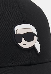 Crna bejzbol kapa sa uzdignutom, višebojnom gumenom oznakom stilizovane face sa belom kosom i tamnim naočarima.