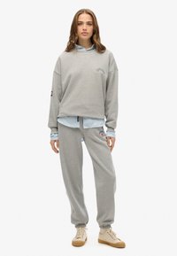 Sudadera gris clara con hombros caídos ligeramente y impresión de logo, superpuesta a una camisa azul clara. Combinada con pantalones de chándal grises a juego.