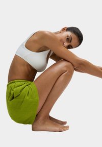 Groene textuurshorts met een gegatherde tailleband en strikaccent, gecombineerd met een witte geribbelde cropped top. Model zit in een squat, waarmee beide items worden getoond.
