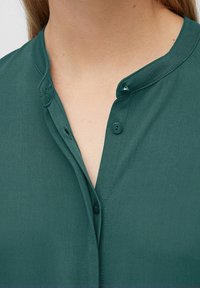 Tummansiniharmaa paita, jossa on hieno kiilto, pyöreä kaulus, kaksi napinläpeä ja sileä pinta. Ei kuvioita tai lisäkoristeita.