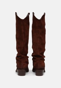 Une paire de bottes de cowboy en daim marron, grandes et désinvoltes, avec des talons en bois épais, vues de l'arrière sur un fond blanc uni.