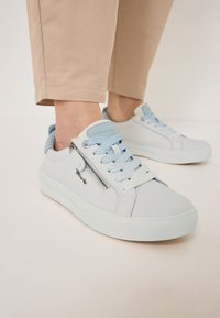 Világosszürke bőr sportcipők sima textúrával. Cipzár díszítéssel, kék fűzőkkel és fehér gumitalppal rendelkezik.