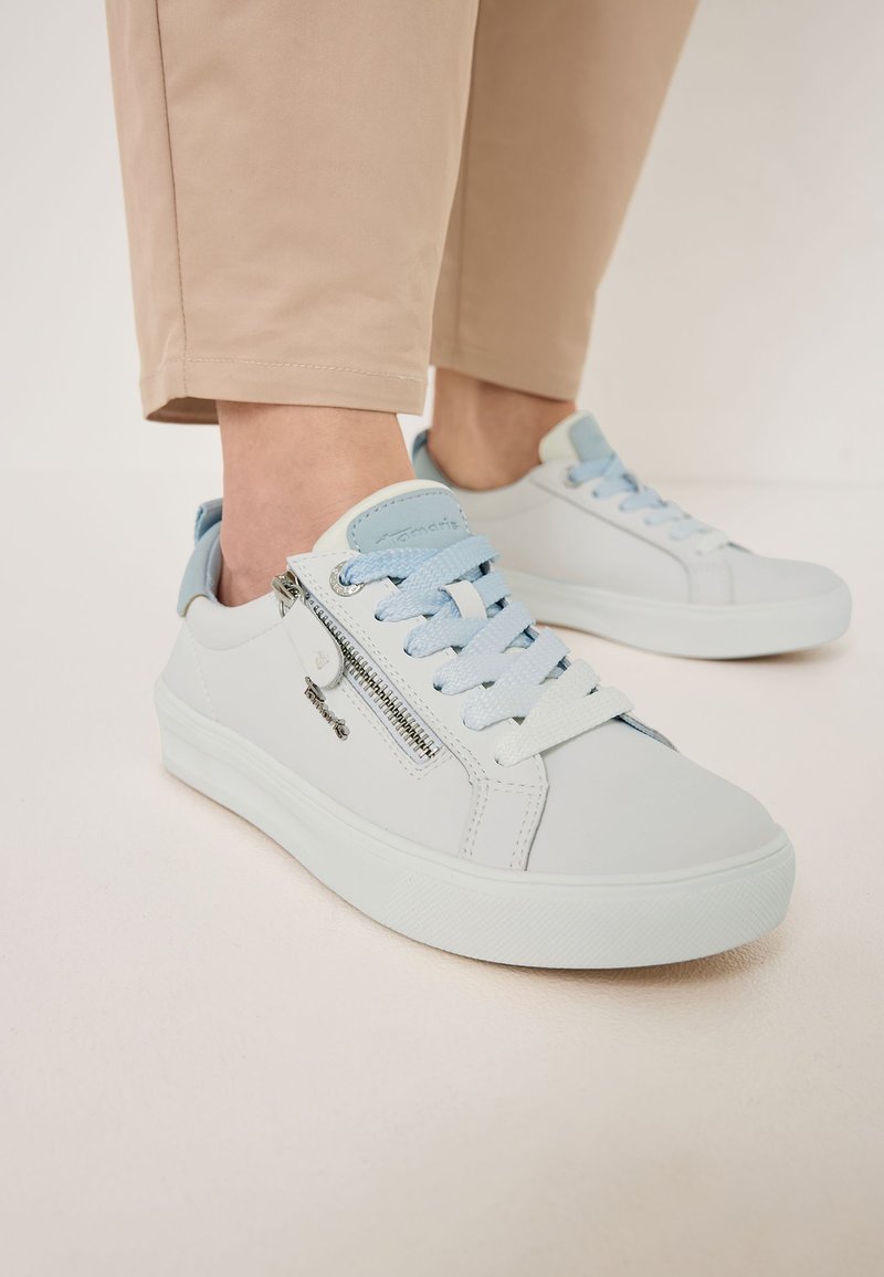 Világosszürke bőr sportcipők sima textúrával. Cipzár díszítéssel, kék fűzőkkel és fehér gumitalppal rendelkezik.