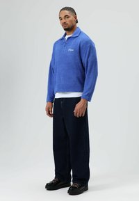 Blauwe fleece trui met kraag, voorzien van een voorzak en logo. Gecombineerd met losse donkere spijkerbroek en zwarte plateau schoenen.