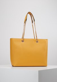 Żółta torba na ramię wykonana z gładkiego materiału, z dwoma uchwytami w formie łańcuszka, metalicznymi akcentami w miejscach mocowania oraz prostokątnym kształtem.