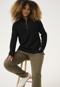 Schwarzer Pullover mit langen Ärmeln und Reißverschlusskragen, strukturierter Stoff; kombiniert mit olivgrünen Hosen und weißen Turnschuhen.