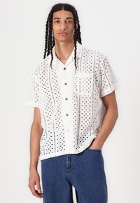 White short-sleeve shirt featuring intricate eyelet patterns, a button-up front, and a single chest pocket; paired with blue jeans.