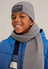 Siva rebrasta pletena kapa z zatemnjenim oznako, v kombinaciji z ujemajočim sivim šalom. Puhasta modra jakna ima zadrgo spredaj in je oblečena čez črtasto pulover.