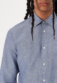 Camicia in chambray blu con colletto, bottoni bianchi e una trama testurizzata. Presenta maniche lunghe e una vestibilità slanciata.