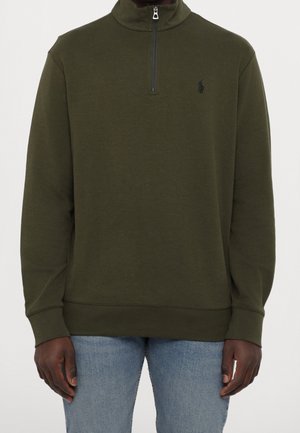 Uomo che indossa una felpa olive verde con zip a quartino, maniche lunghe e piccolo logo nero ricamato, abbinata a jeans azzurri.