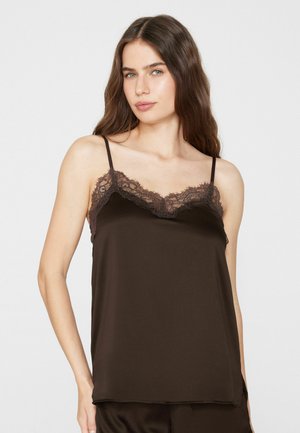 Junge Frau mit langen braunen Haaren, die ein dunkelbraunes Satin-Camisole mit Spitzenbesatz trägt und vor einem schlichten weißen Hintergrund steht.