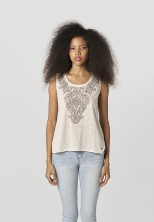 Bež brezrokavnik z zapletenimi temnimi okraski, mehka tkanina, ohlapen kroj in zaobljen izrez, kombiniran z svetlo modrimi ozkimi kavbojkami.