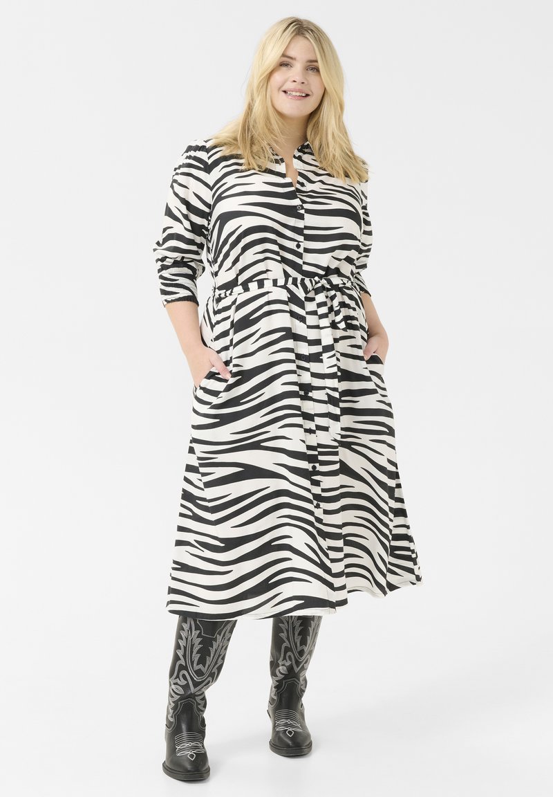 Abito lungo con bottoni, caratterizzato da una stampa zebra bianca e nera, dotato di una cintura, due tasche laterali e maniche lunghe. Indossato con stivali neri ricamati.