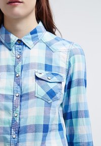 Camicia a quadri blu chiaro con accenti blu scuro. Presenta un colletto, maniche lunghe, due tasche sul petto e un orlo arrotondato.