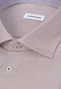 Gros plan sur un col de chemise marron clair texturé avec des boutons blancs et une étiquette de marque Seidensticker à l'intérieur.