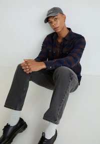 Model wearing a blue plaid flannel shirt over a brown top, gray jeans, black shoes with yellow accents, and a gray cap with text.