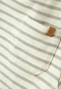 Striped cotton fabric in beige and gray, featuring a chest pocket and a small leather tag with brand name. Soft texture with visible stitching.