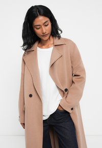Lungo cappotto beige in lana con colletto a rever, bottoni neri doppi e tasche laterali, indossato sopra una camicia bianca e jeans scuri.