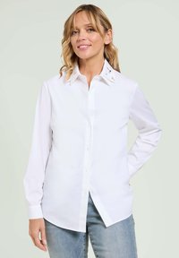 Una camicia bianca con bottoni, realizzata in un tessuto liscio, con dettagli decorativi sul colletto. Maniche lunghe e design dal fit rilassato.