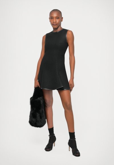 Rochie neagră fără mâneci, cu corset ajustat și fustă conică, având accente lucioase și o textură netedă. Modelul ține o geantă din blană artificială.