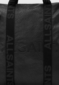Bolso tote de cuero negro con superficie texturizada, marca grabada y asas de tela negra con acentos de logo impresos.