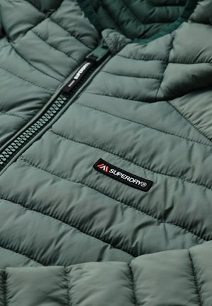 Veste matelassée verte avec une fermeture éclair noire et une étiquette de marque Superdry® à l'avant, montrant des coutures détaillées et la texture du tissu.