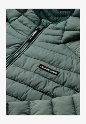 Groene gewatteerde jas met zwarte ritssluiting en Superdry® merklabel aan de voorkant, met gedetailleerde stiksels en stoftextuur.