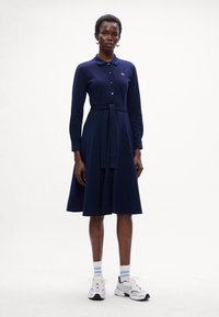 Abito polo blu navy con maniche lunghe, chiusura a bottoni e vita legata. Il tessuto appare morbido, con una silhouette A-line rilassata. Abbinato a sneakers bianche.