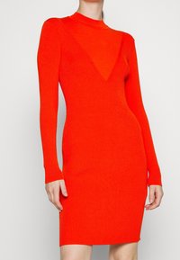 Robe en maille de couleur orange vif avec une silhouette ajustée, des manches longues et un décolleté plongeant en V. Présente une texture côtelée sur tout le tissu.