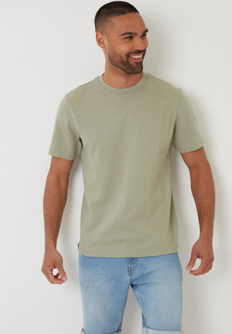 Uśmiechnięty mężczyzna z krótkimi włosami, ubrany w jasnoszary zielony T-shirt z krótkim rękawem i jasnoniebieskie szorty dżinsowe, stojący przy białej ścianie.