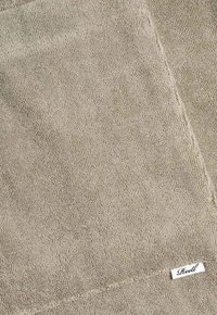 Handtuch aus strukturiertem Stoff in hellgrau-beige, mit sichtbarem gesticktem Rand und einem kleinen Markenetikett in der unteren Ecke.