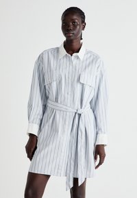 Svītrainā krekla kleita gaiši zilā un baltā krāsā, ar jostu viduklī, divām priekšējām kabatām, pogu aizdari un kontrastējošu baltu apkakli.