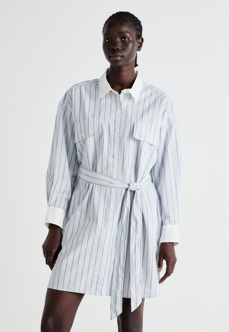 Svītrainā krekla kleita gaiši zilā un baltā krāsā, ar jostu viduklī, divām priekšējām kabatām, pogu aizdari un kontrastējošu baltu apkakli.