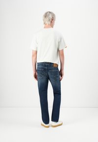 Person with short blond hair standing, wearing a white t-shirt, dark blue jeans, and white sneakers against a plain white background.